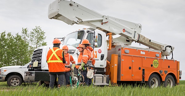 Four SaskPower employees standing in a circle talking in front of a SaskPower bucket truck wearing orange hard hats and in power line technician gear. 