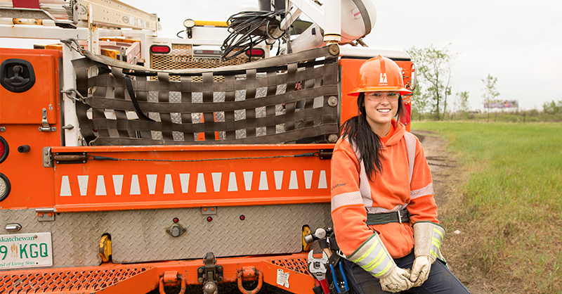 A female sitting on the back of a SaskPower trucking and smiling while wearing an orange sweater, SaskPower hard hat, safety goggles and gloves.