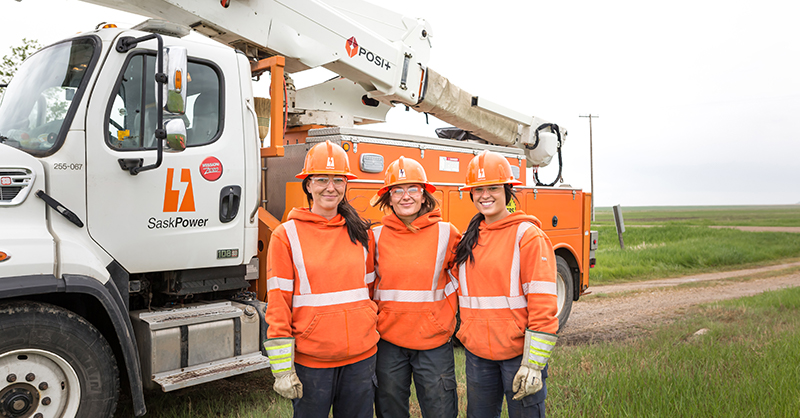 Three females standing in front of a SaskPower bucket truck wearing orange hard hats and sweaters. 