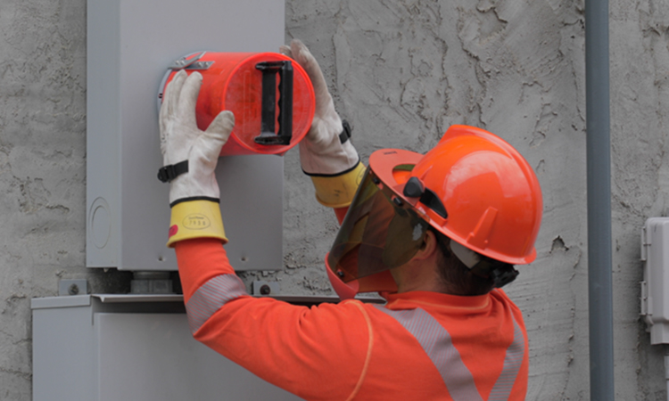 SaskPower technician wearing an orange hard hat and shirt installing a smart meter on a home. 