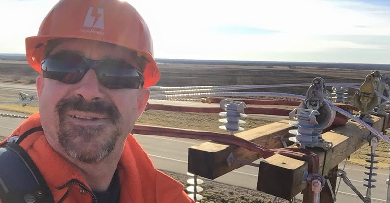 A male wearing an orange SaskPower hard hat with a power pole in the background.