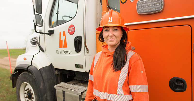 Breanne standing in front of a SaskPower bucket truck, smiling and wearing an orange hard hat and sweater.
