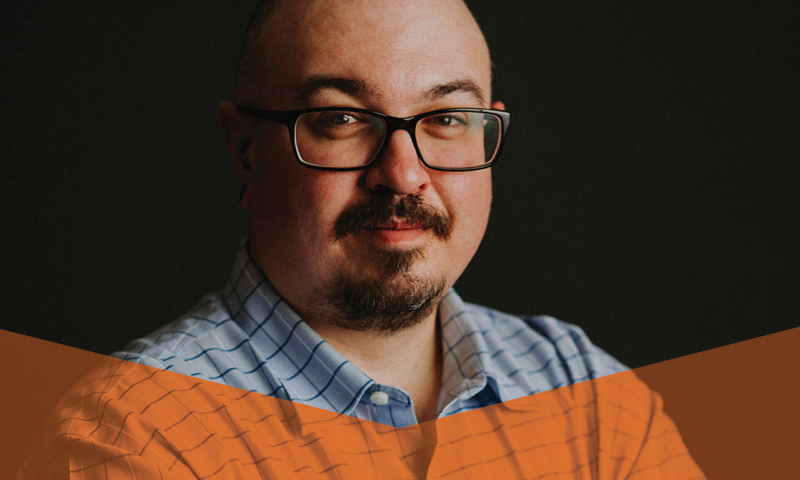 A man smiling at the camera wearing a blue shirt and glasses.