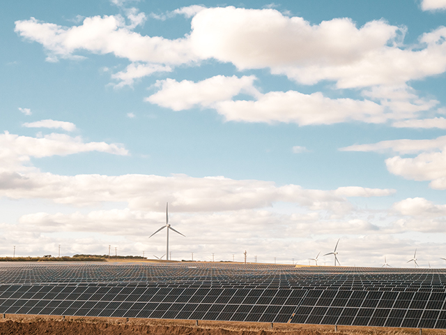 Rows of solar panels with wind turbines behind in the distance.